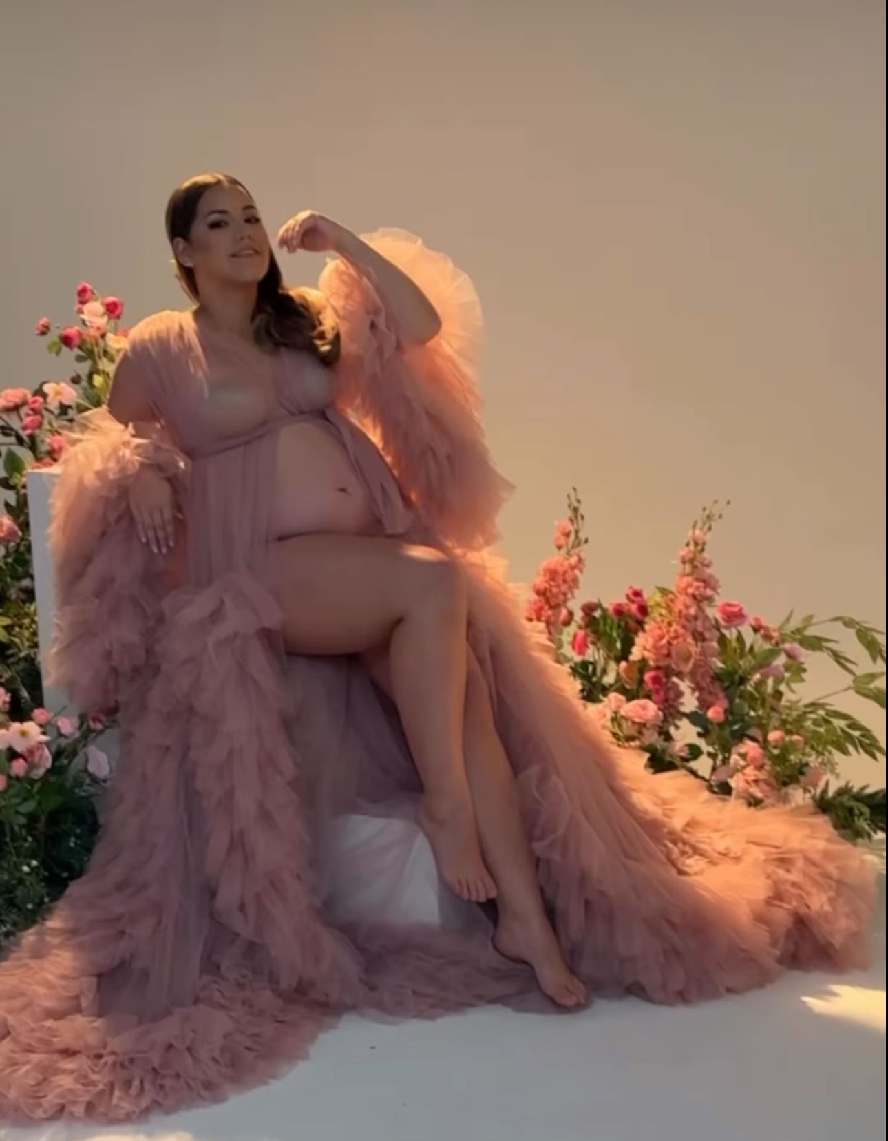 Pregnant woman posing in a flowing pink tulle maternity robe surrounded by flowers.