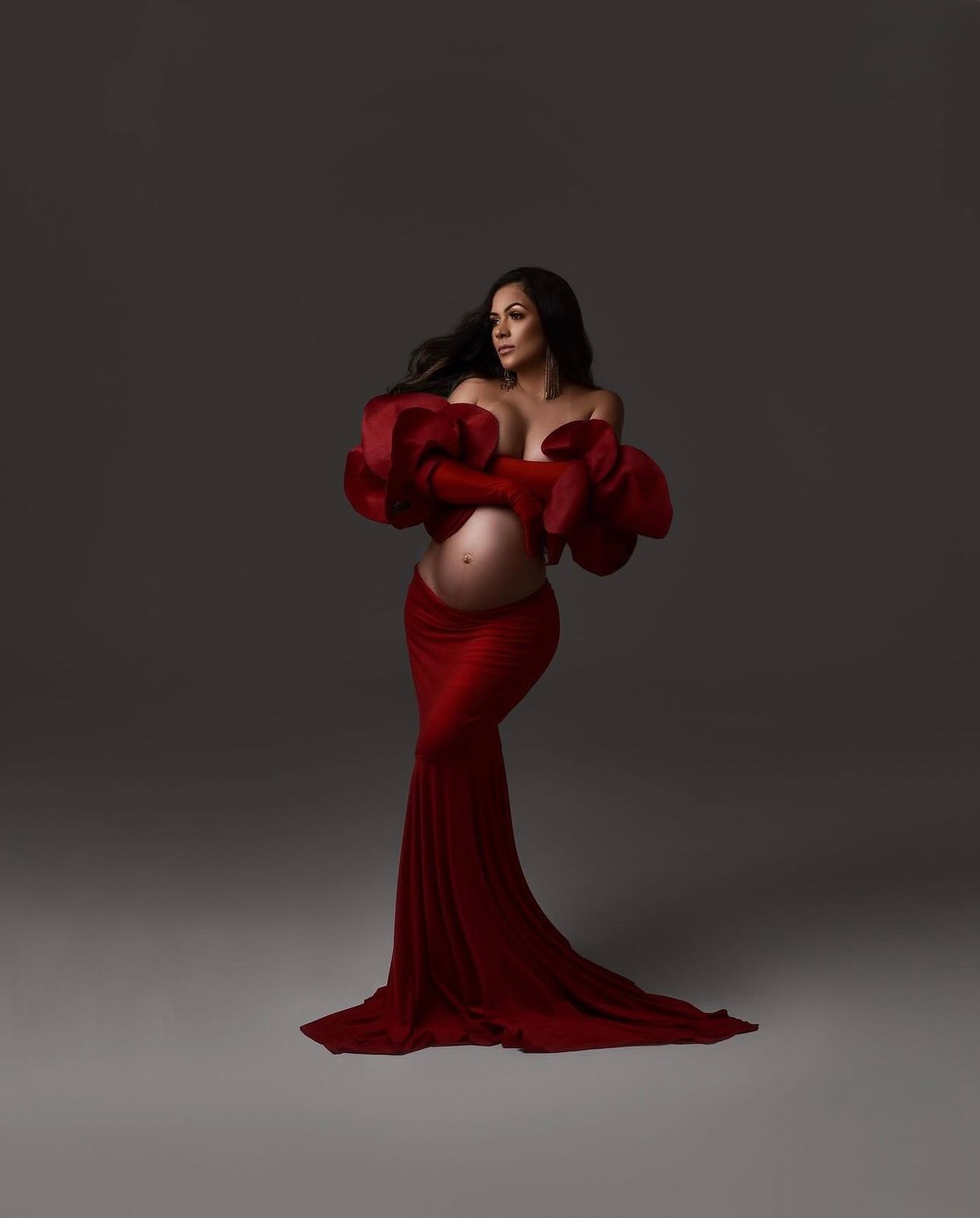 Pregnant woman in elegant red off-shoulder gown with dramatic sleeves and train poses gracefully in studio maternity photo.