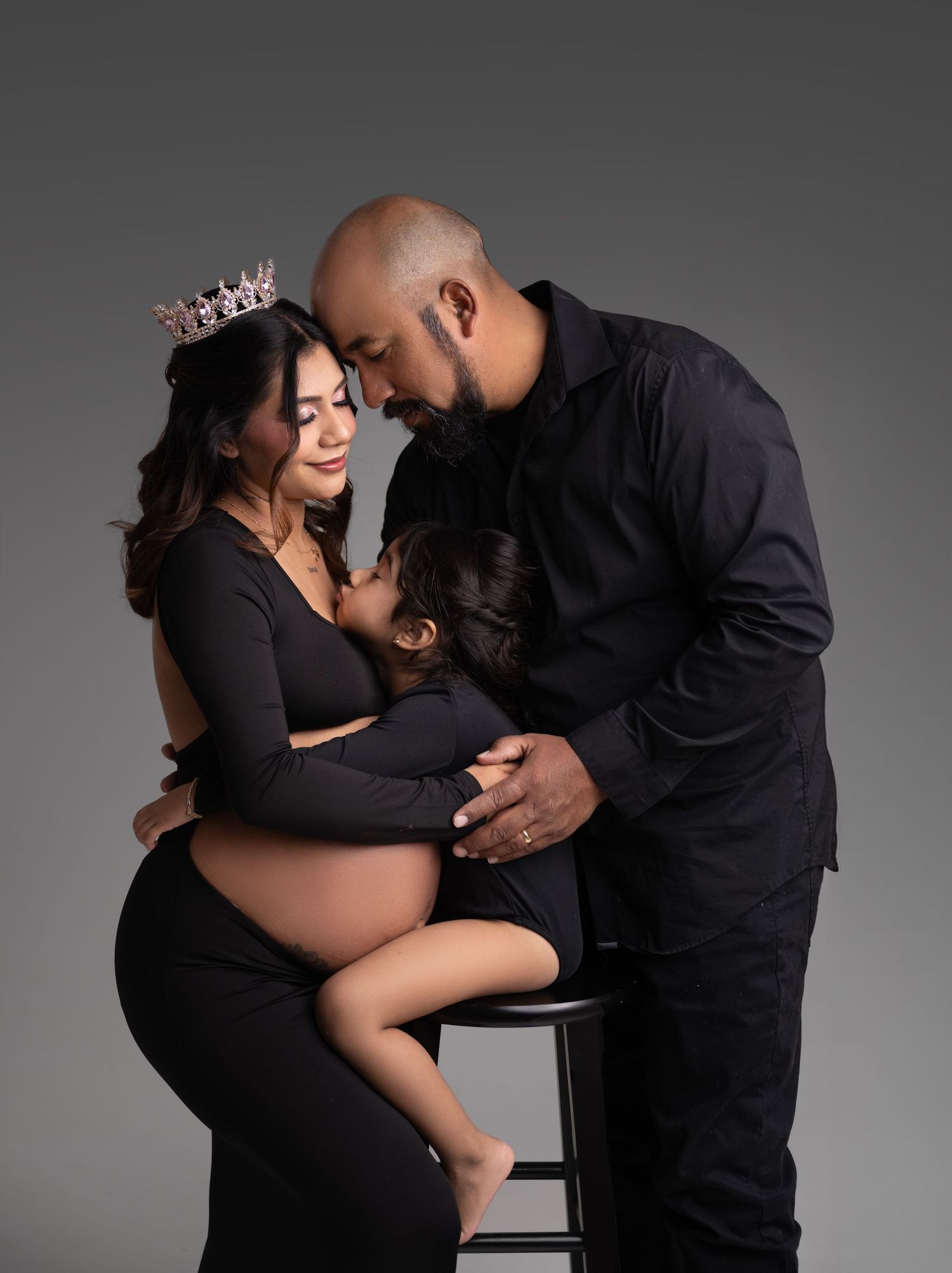 A romantic maternity photoshoot features a couple in black outfits embracing against a gray studio backdrop.