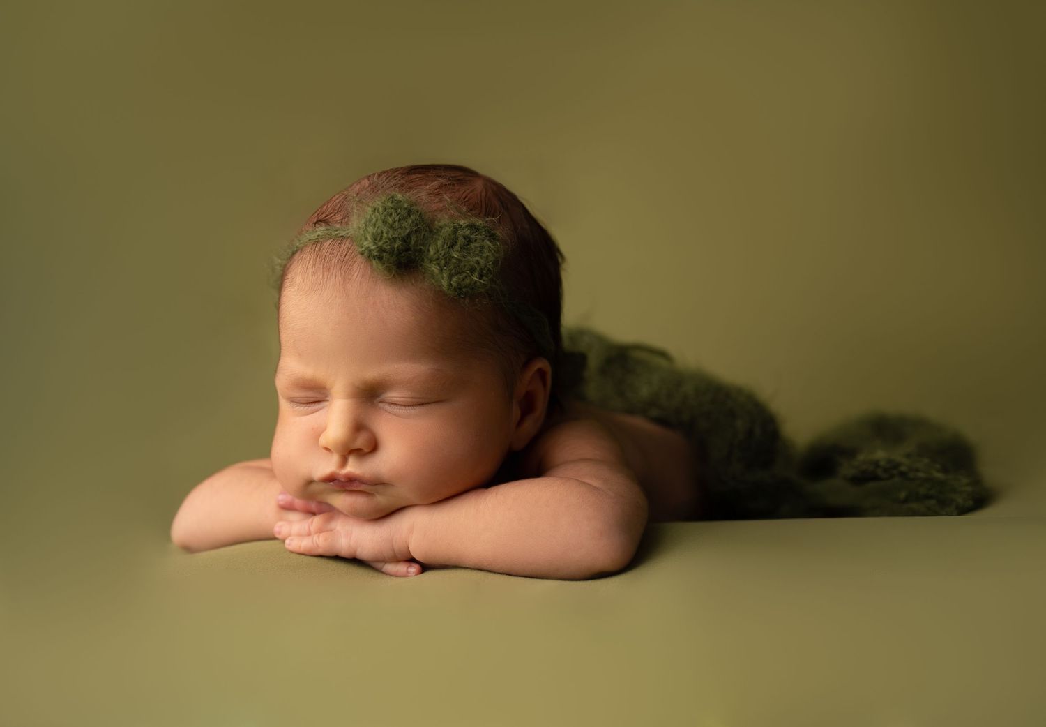 Peaceful newborn portrait with green accessories against olive backdrop.