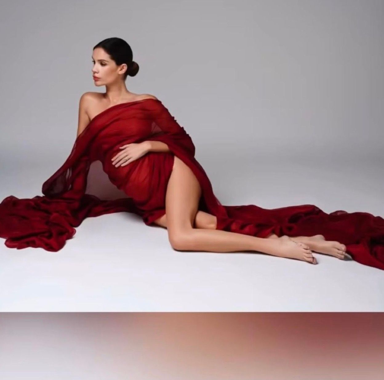 Pregnant woman in elegant red flowing maternity gown posed gracefully on white floor for professional photo shoot.
