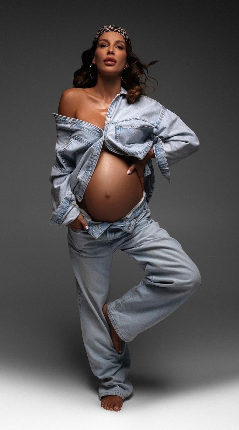 Pregnant woman posing in off-shoulder denim jacket and wide-leg jeans, showcasing baby bump in stylish maternity photoshoot.