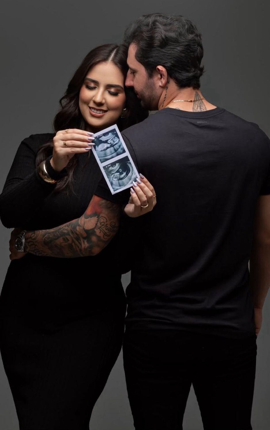 Couple in black outfits sharing a joyful moment while holding ultrasound photos, announcing their pregnancy.