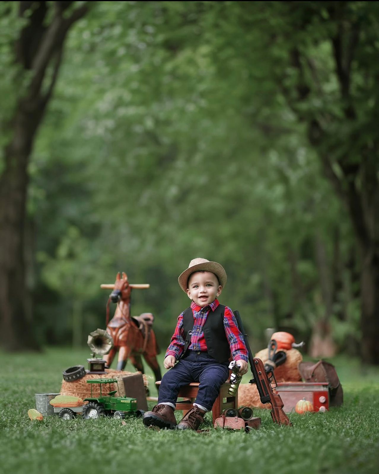 A toddler boy in a plaid shirt and hat sits outdoors surrounded by vintage toy horses and wooden props on green grass.