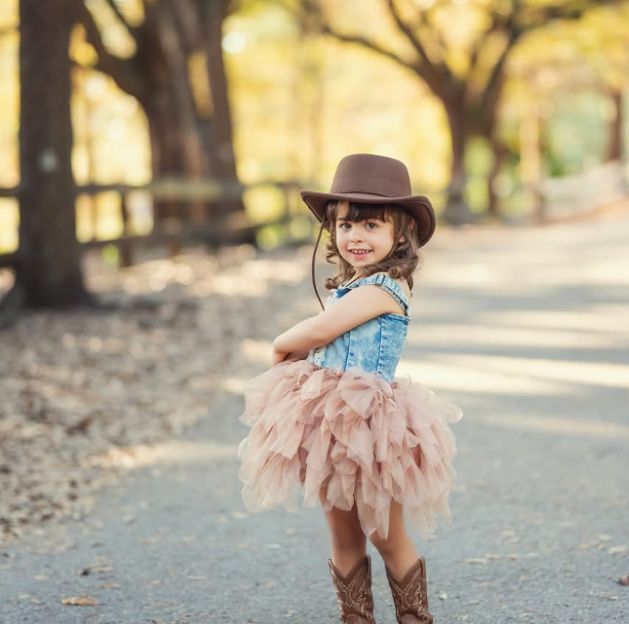 A young girl in a denim top, pink tutu, cowboy hat, and boots poses on a tree-lined path.