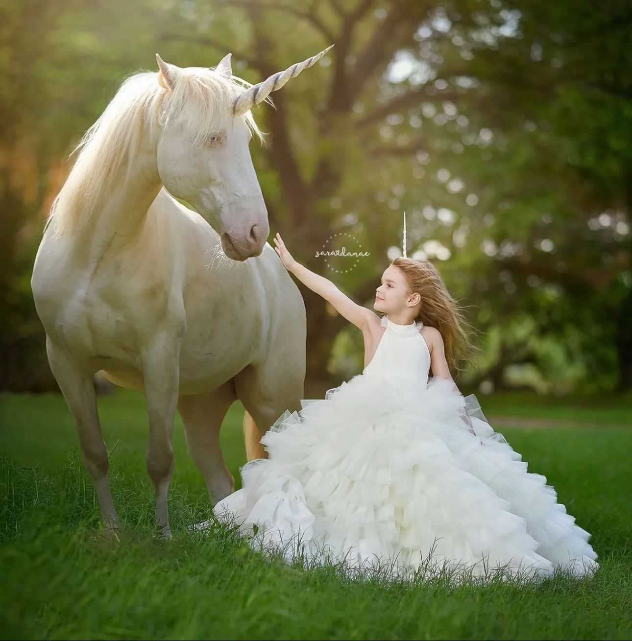 A young girl in a white ball gown dress reaches out to touch a white unicorn on a lush green lawn.