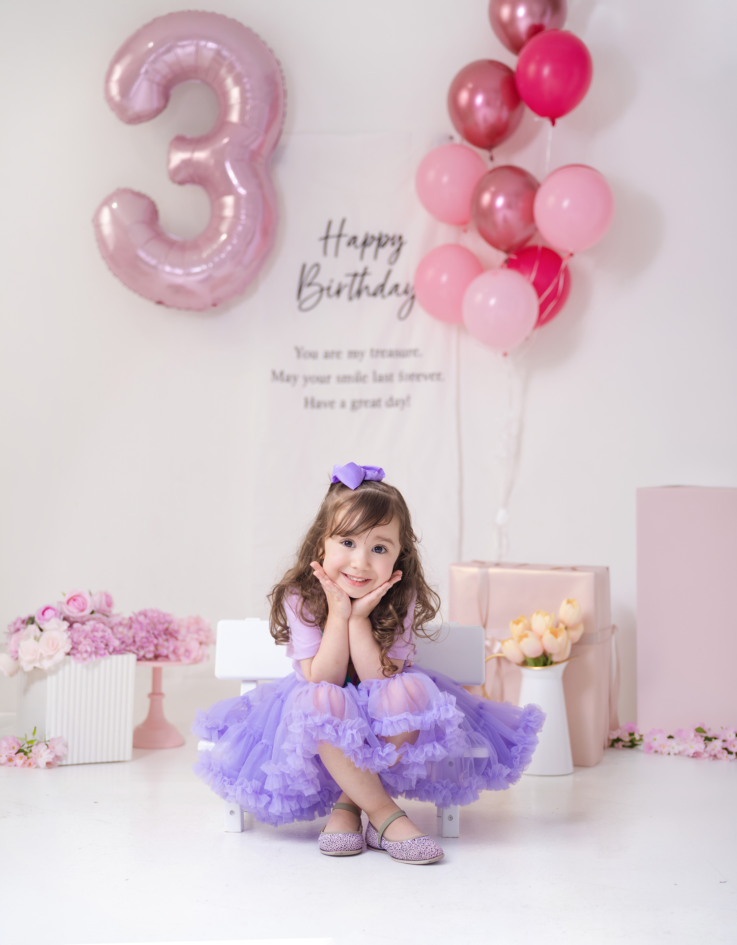 A smiling girl in a purple tutu dress poses at her 3rd birthday party with pink balloons and Happy Birthday backdrop.