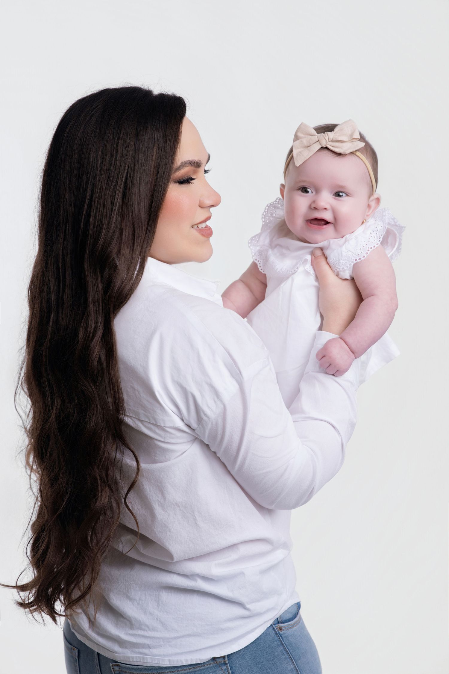 Young mother with long dark hair holding smiling baby girl in white dress with bow headband.