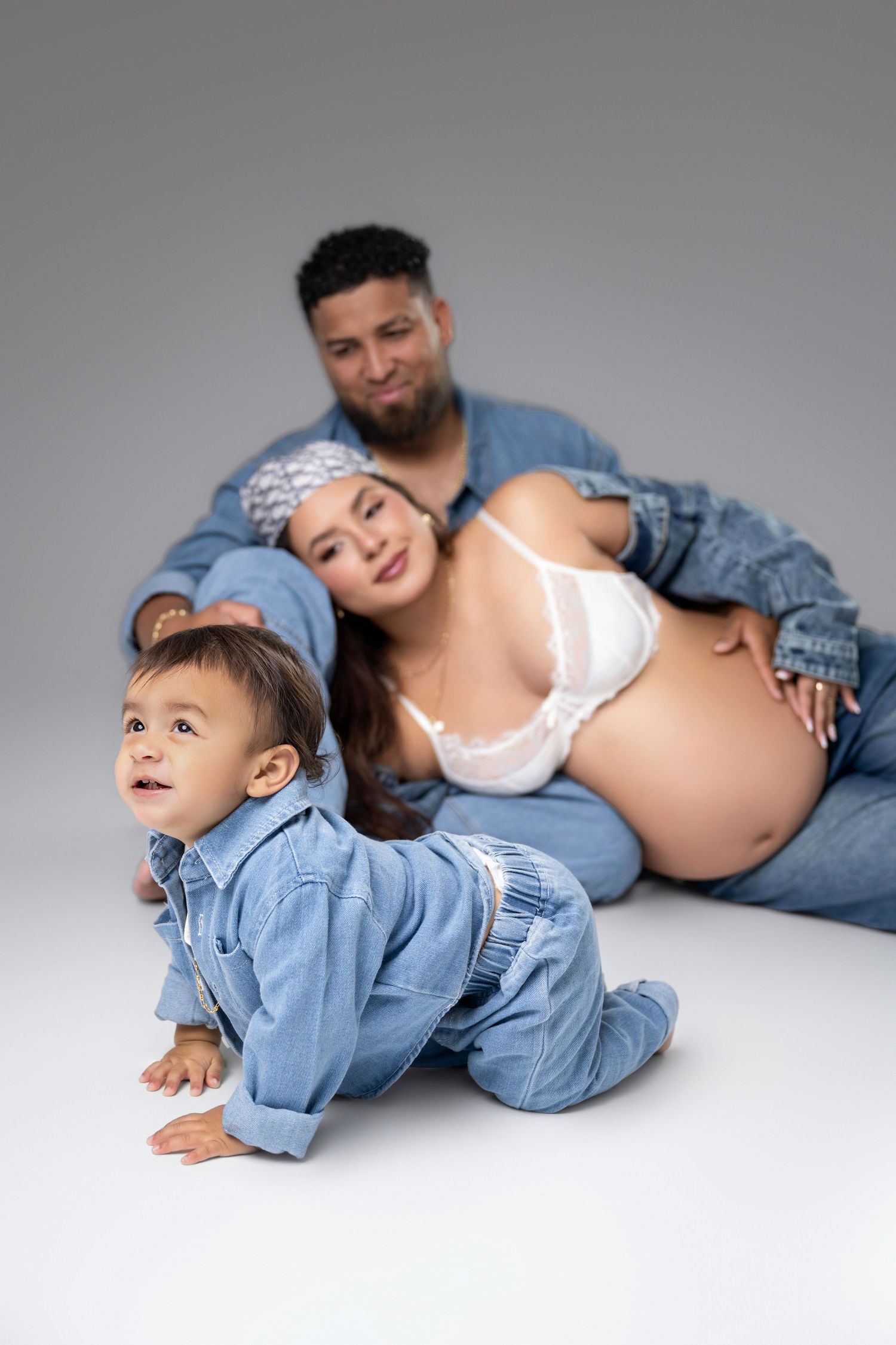 A pregnant woman in white lace bra and jeans sits with her partner while their toddler crawls in the foreground.