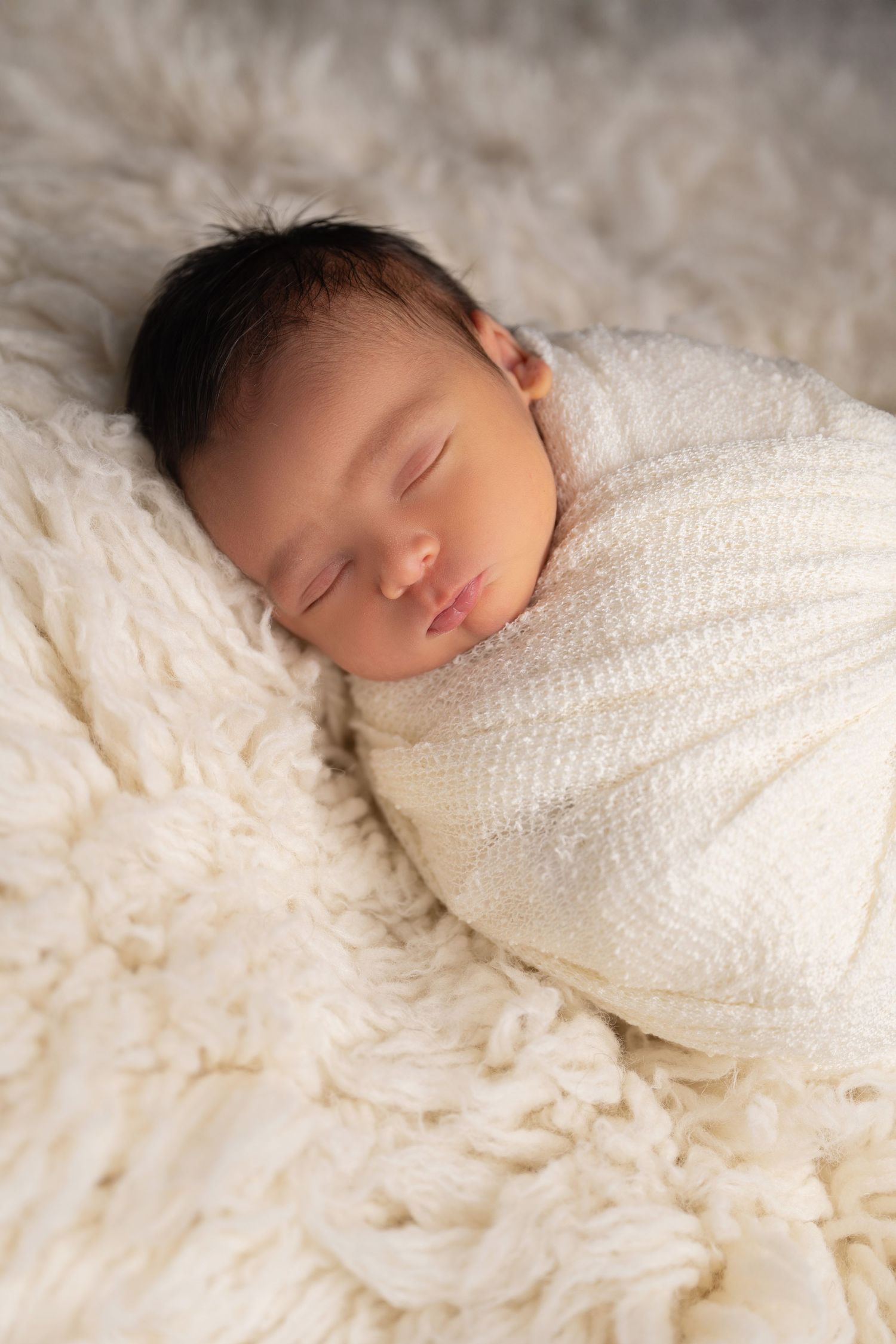 Peaceful sleeping newborn wrapped in white knit blanket on fuzzy cream backdrop.