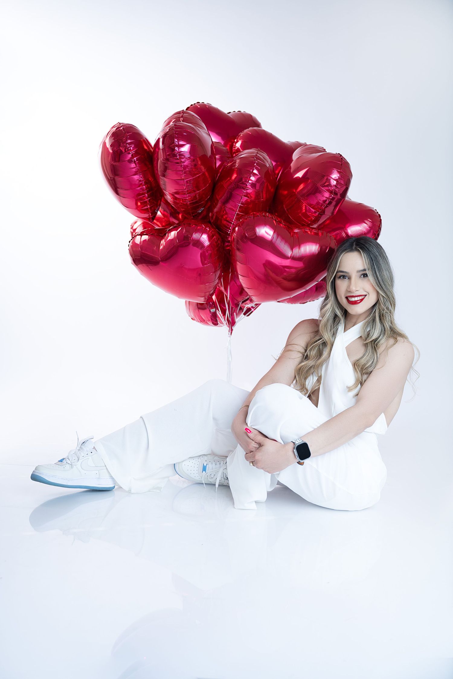 Woman in white outfit sitting on floor with red heart-shaped balloons against white background.