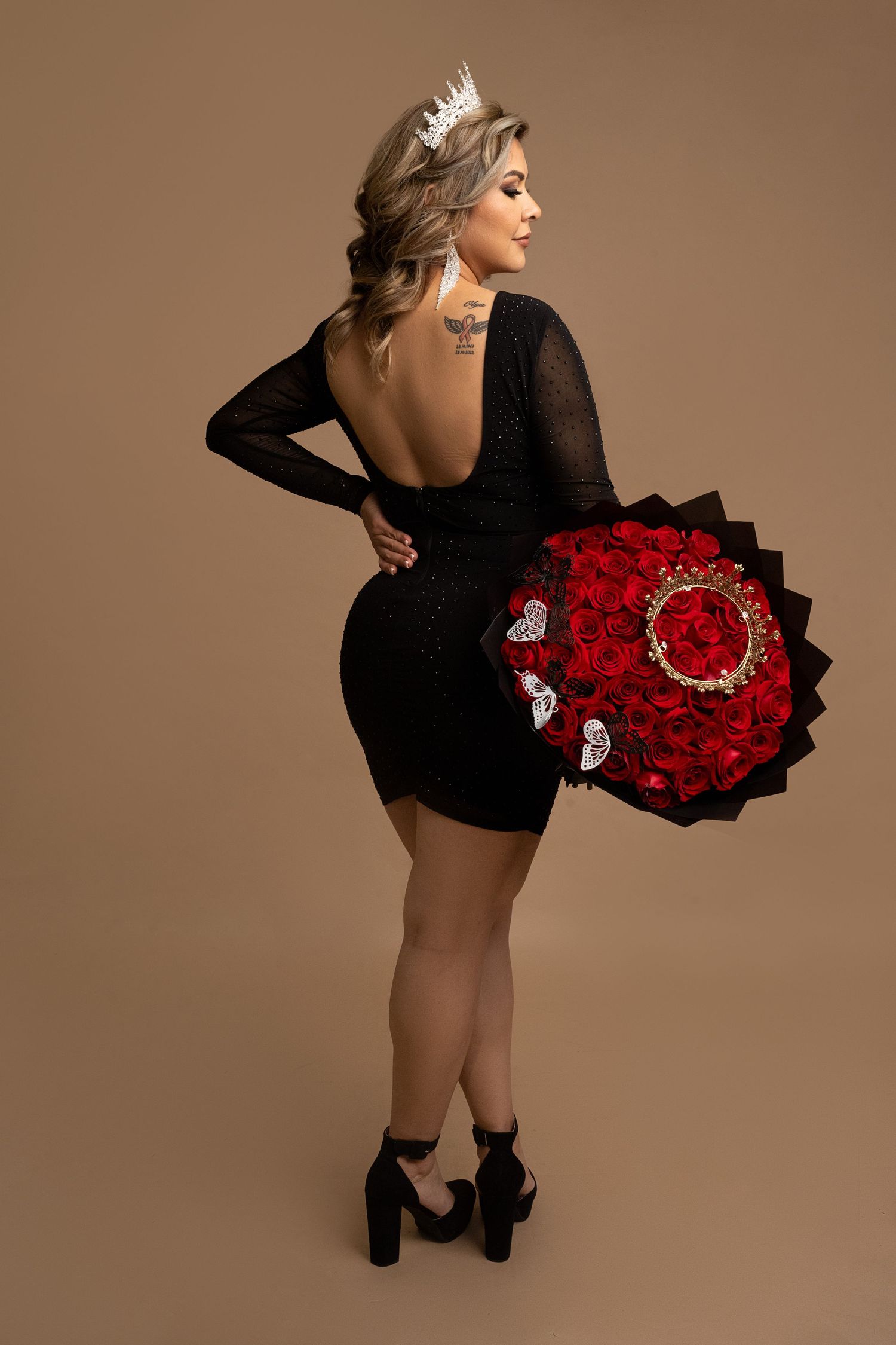Glamorous photoshoot featuring a black backless dress and dramatic red rose bouquet against brown background.