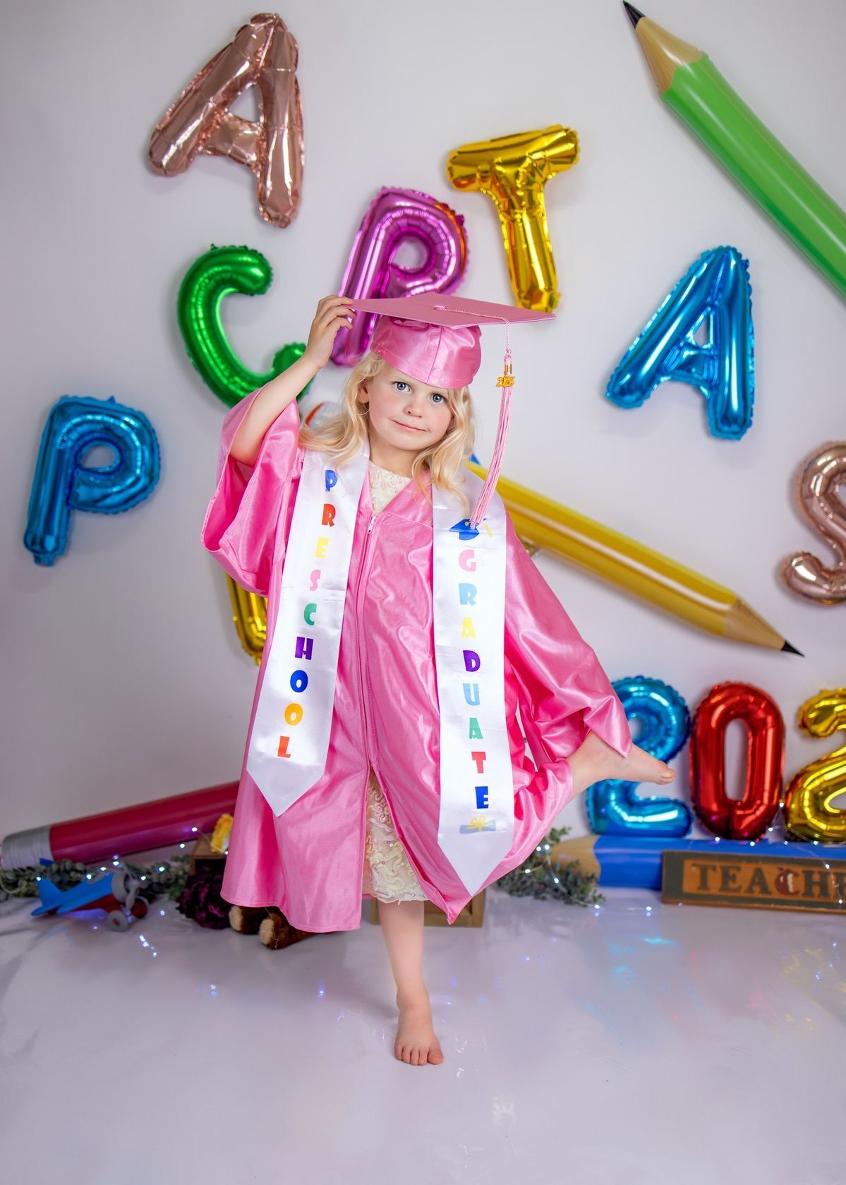 Young girl in pink graduation cap and gown poses with colorful letter balloons and pencil decorations at preschool graduation.