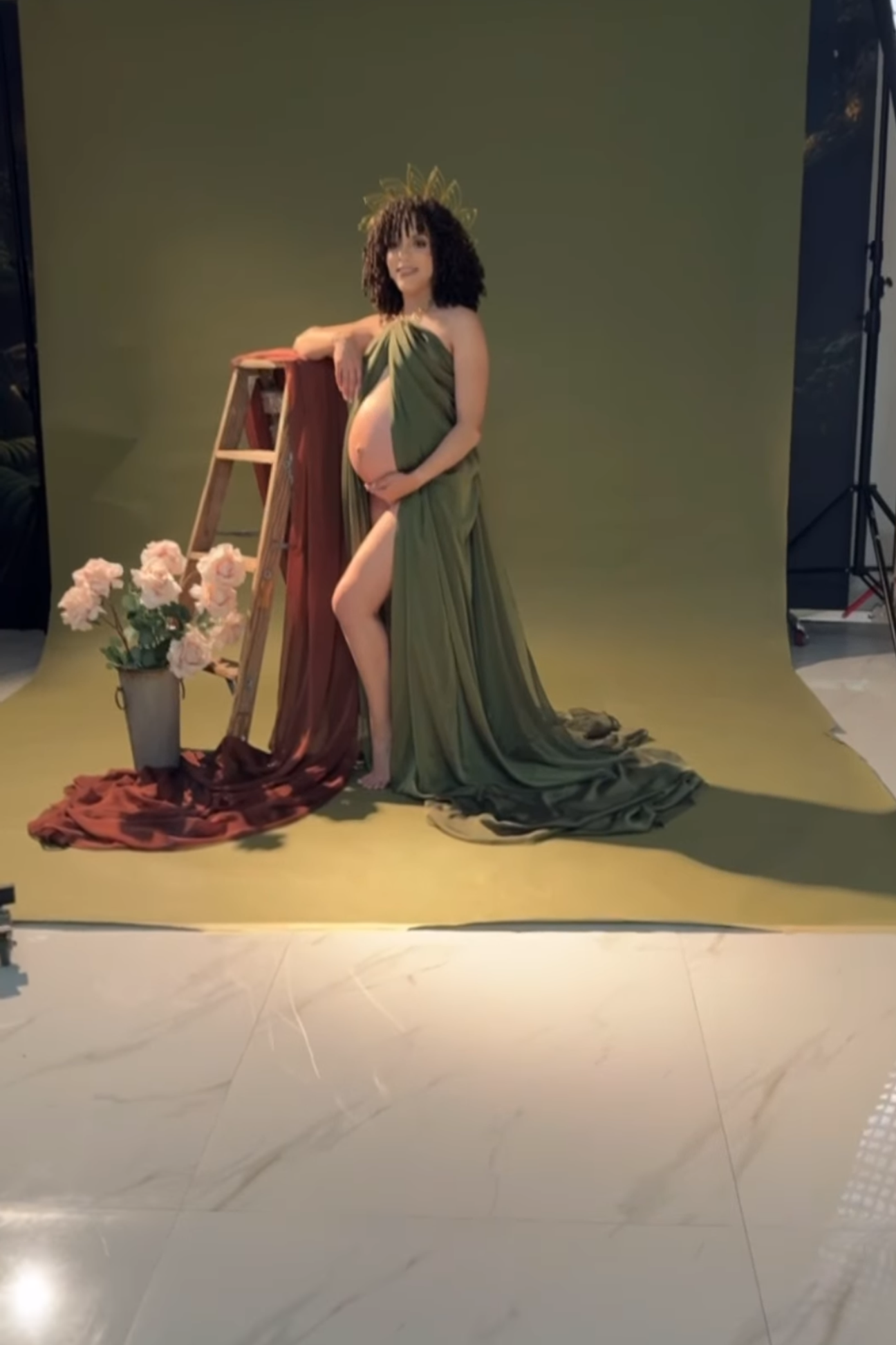 Pregnant woman in green draped gown with floral crown poses beside ladder and pink flowers in photography studio.