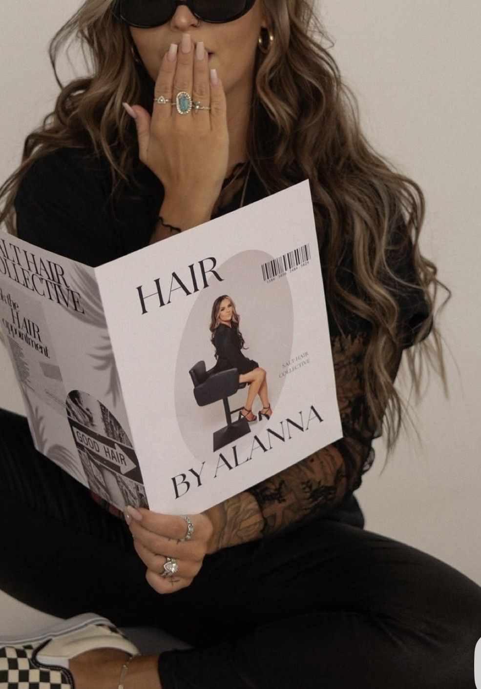 Woman in black outfit holding a Hair by Alanna magazine, showcasing long wavy hair and stylish rings.
