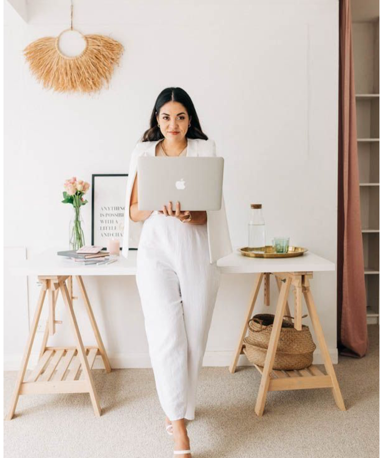 Woman in white blazer and pants holding MacBook laptop standing between two wooden trestle desks in bright home office.