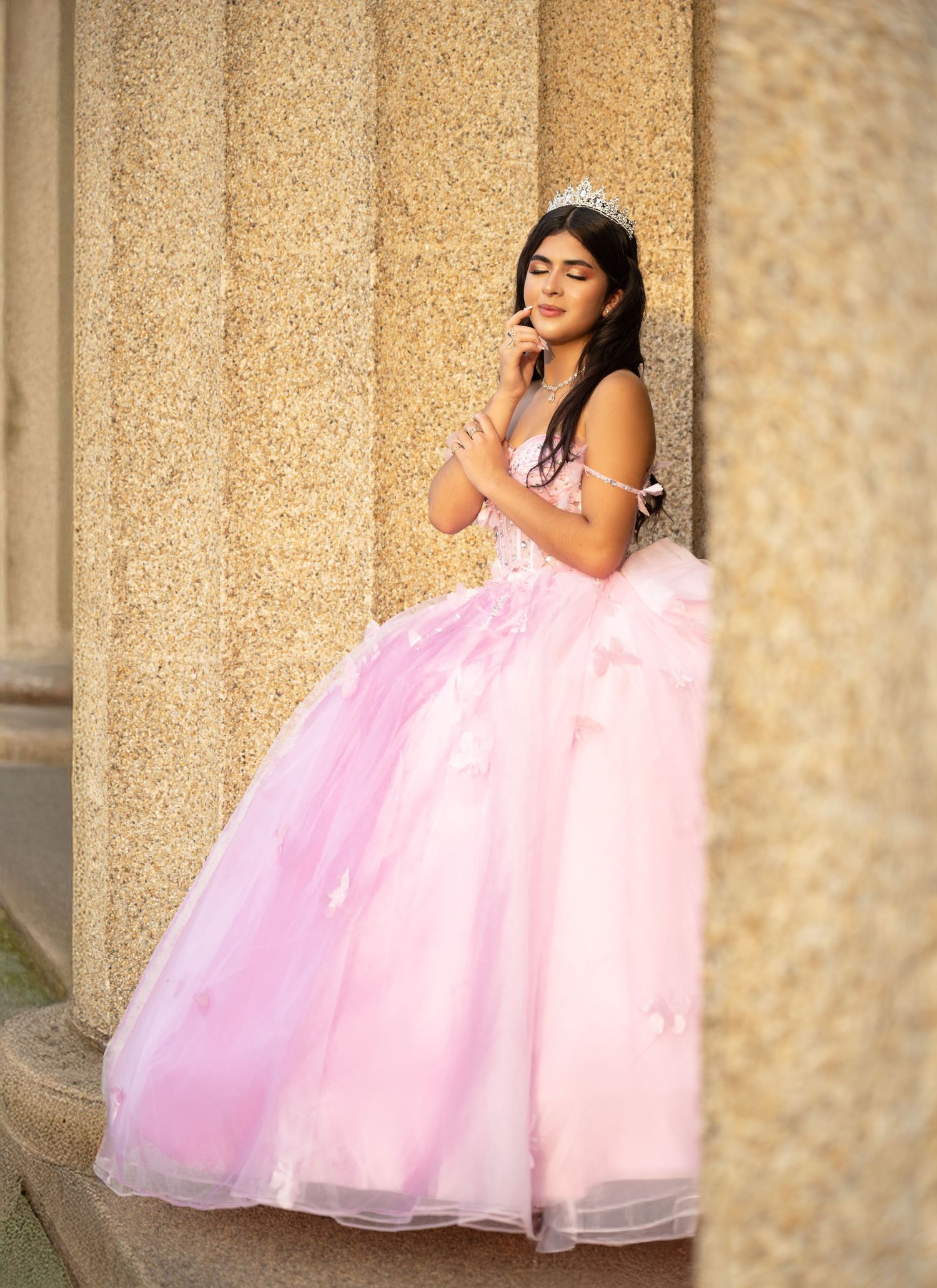 Young girl in pink ball gown and tiara poses gracefully between stone columns for quinceañera portrait.