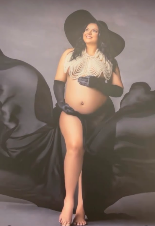 A figure in a dramatic black hat and pearls poses against a dark background in an artistic maternity photoshoot.