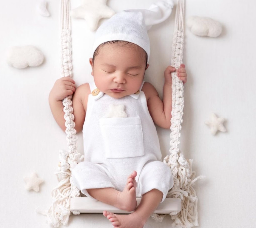 Newborn baby in white outfit resting peacefully in macrame hanging basket.