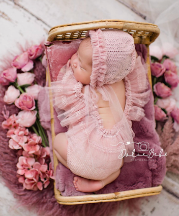 Delicate pink baby booties with ruffles surrounded by soft pink flowers and petals on a light surface.