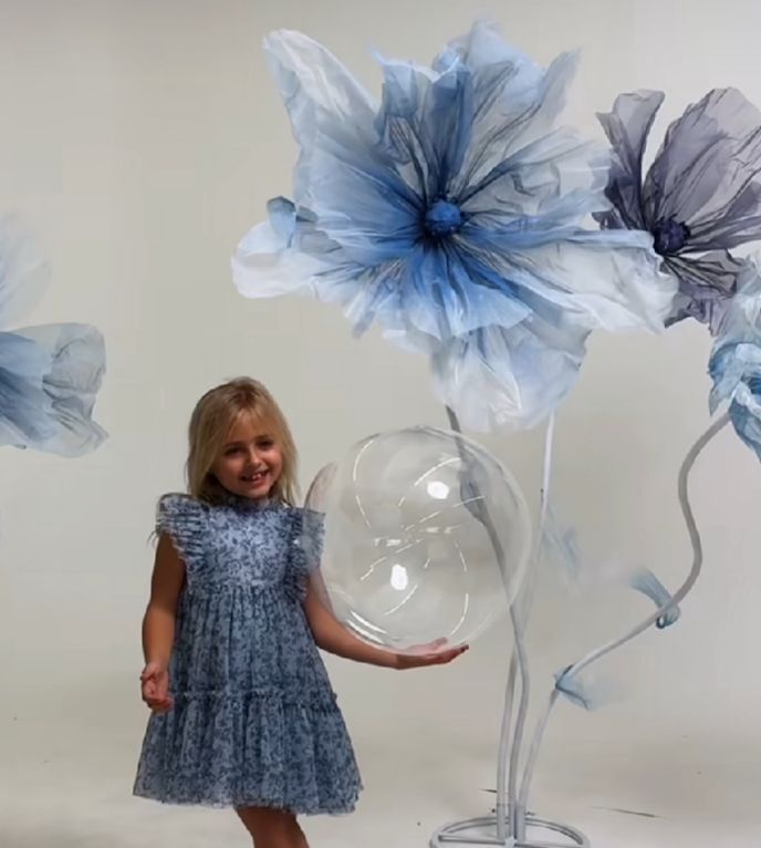 A child in a blue ruffled dress stands beside large decorative blue floral wall art and an oversized wine glass.