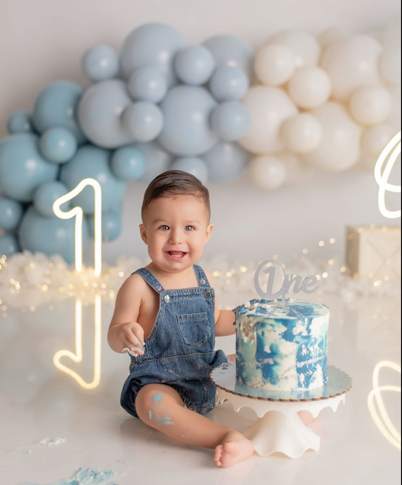 A baby in denim overalls sits near a blue and white birthday cake with balloon decorations in the background.