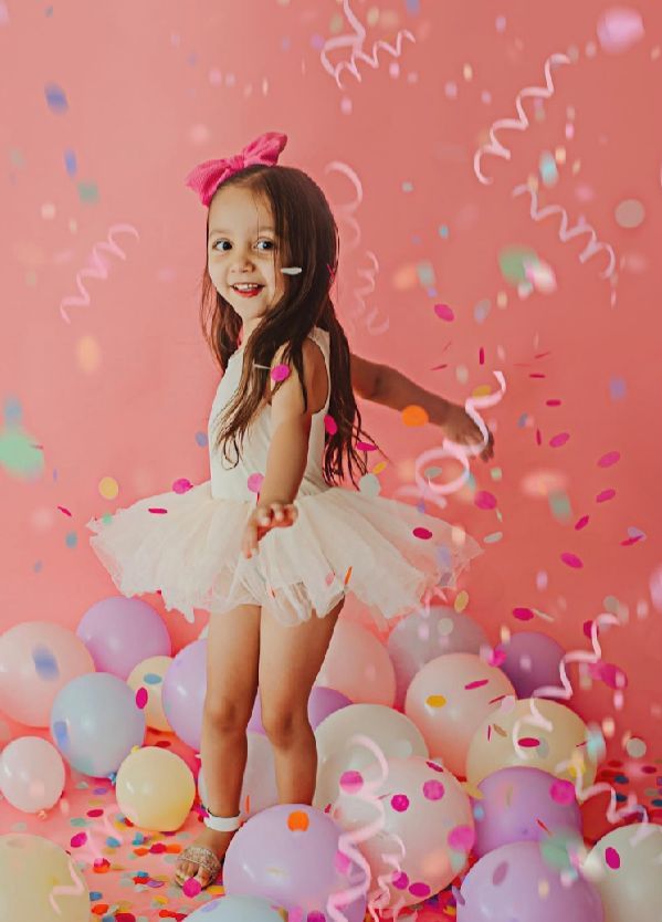 A child in a white tutu dress jumps playfully among pink balloons and colorful confetti on a pink background.