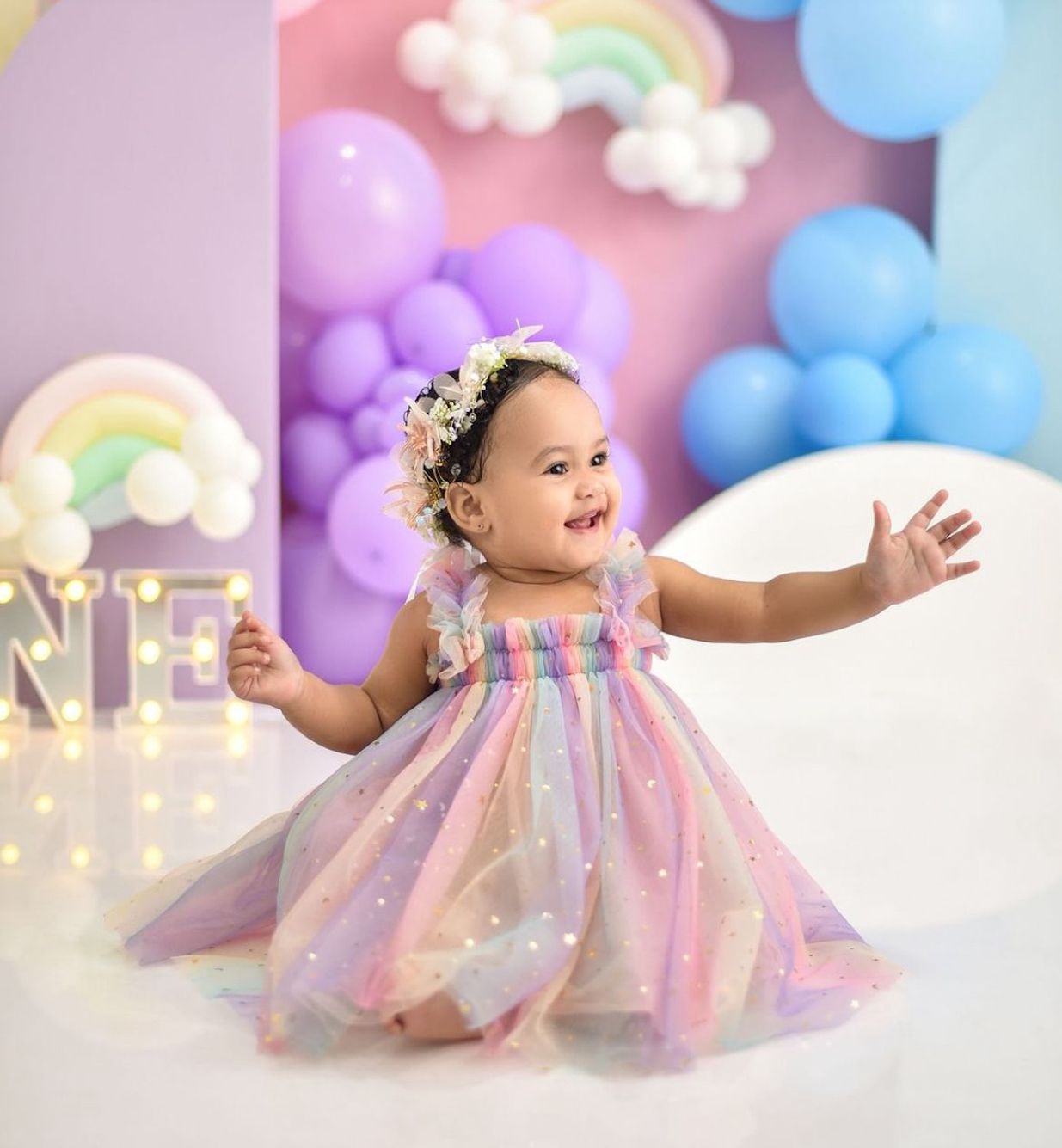 A baby in a rainbow tulle dress smiles and plays during a dreamy pastel balloon-filled cake smash photo session.