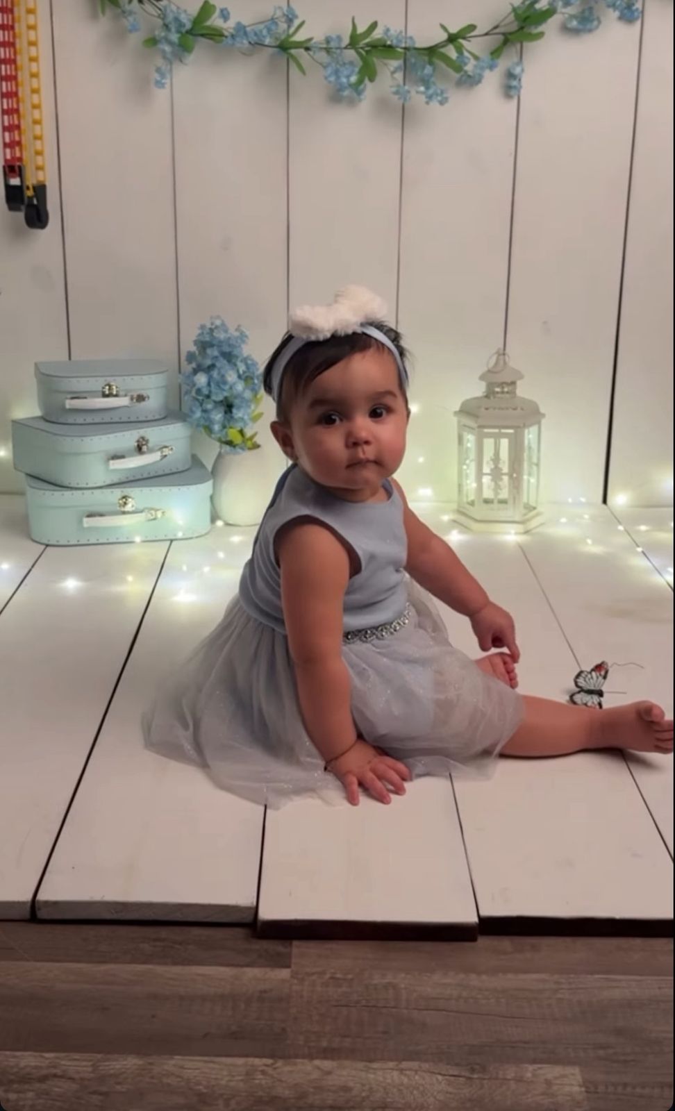 A baby in a gray and white tulle dress sits on a white wooden floor with vintage suitcases and lantern decor.