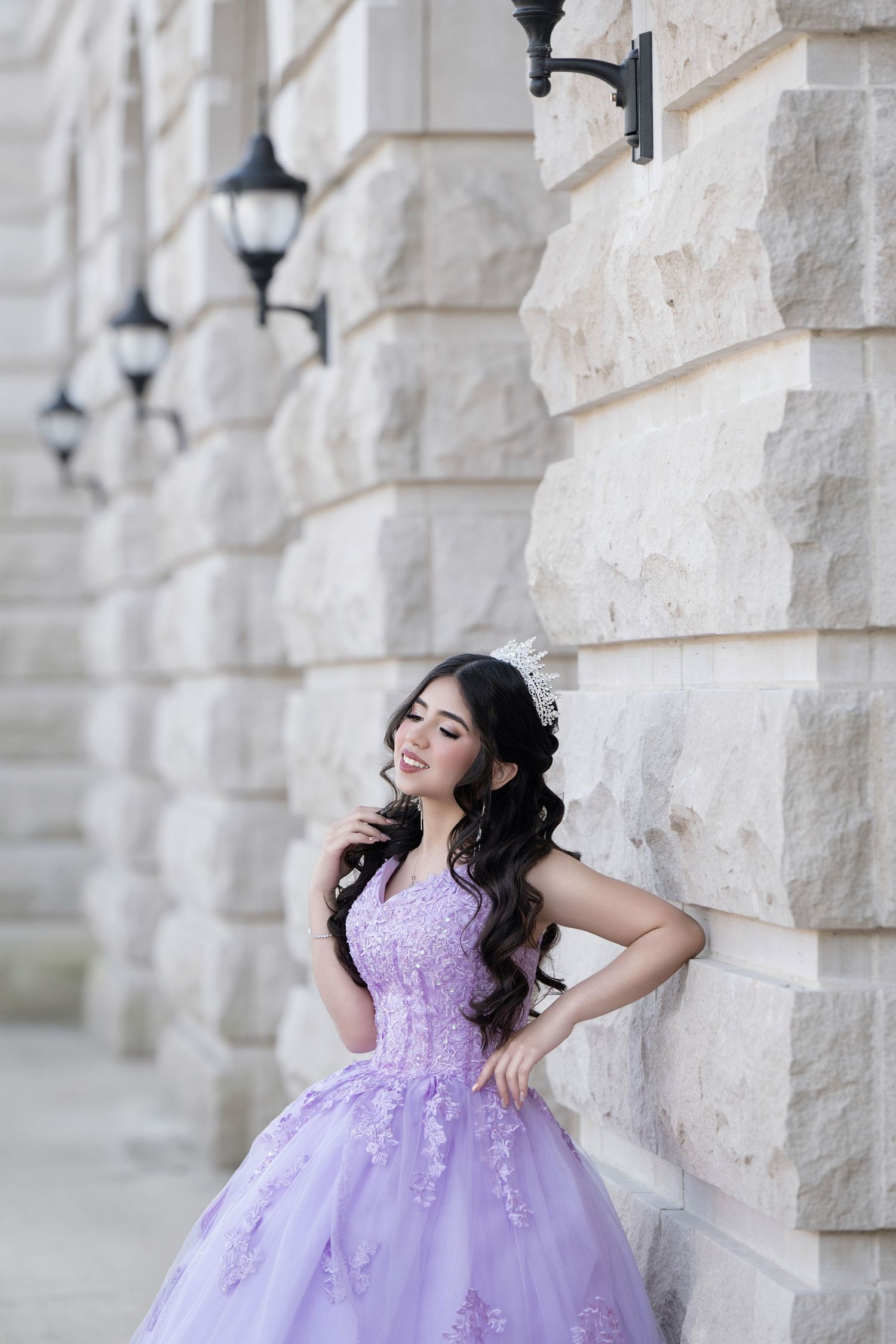 Young woman in lavender ball gown with floral lace and tiara poses gracefully against stone building exterior.