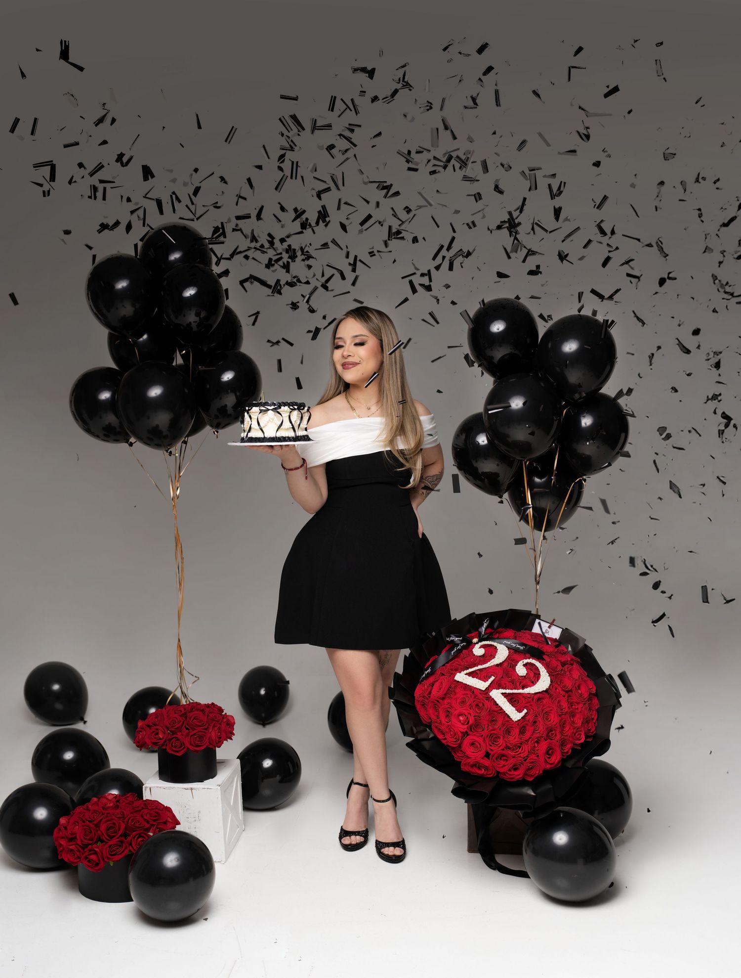 A black little dress paired with red roses and black balloons creates a dramatic fashion scene against a white backdrop.