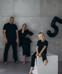 A family of three wearing matching black outfits poses against a concrete wall with minimalist styling.