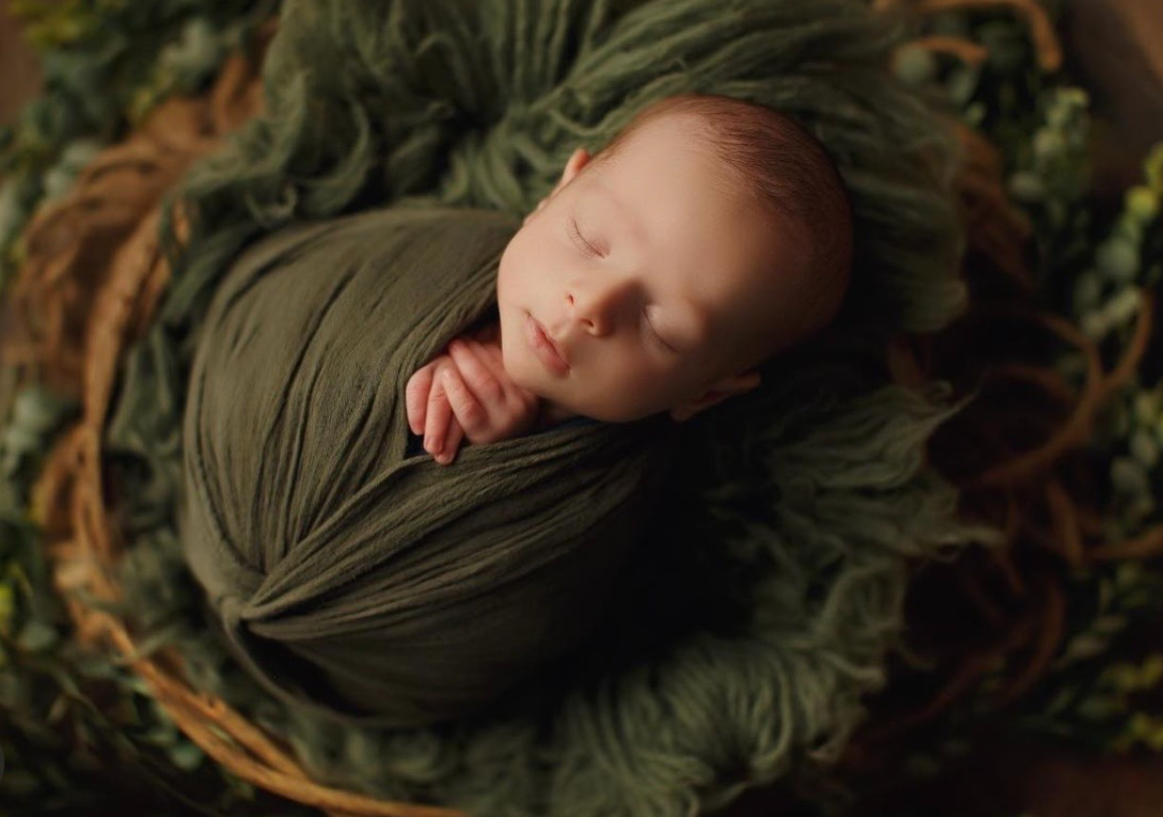 A sleeping newborn wrapped in olive green fabric nestles in a rustic wooden basket surrounded by autumn foliage.