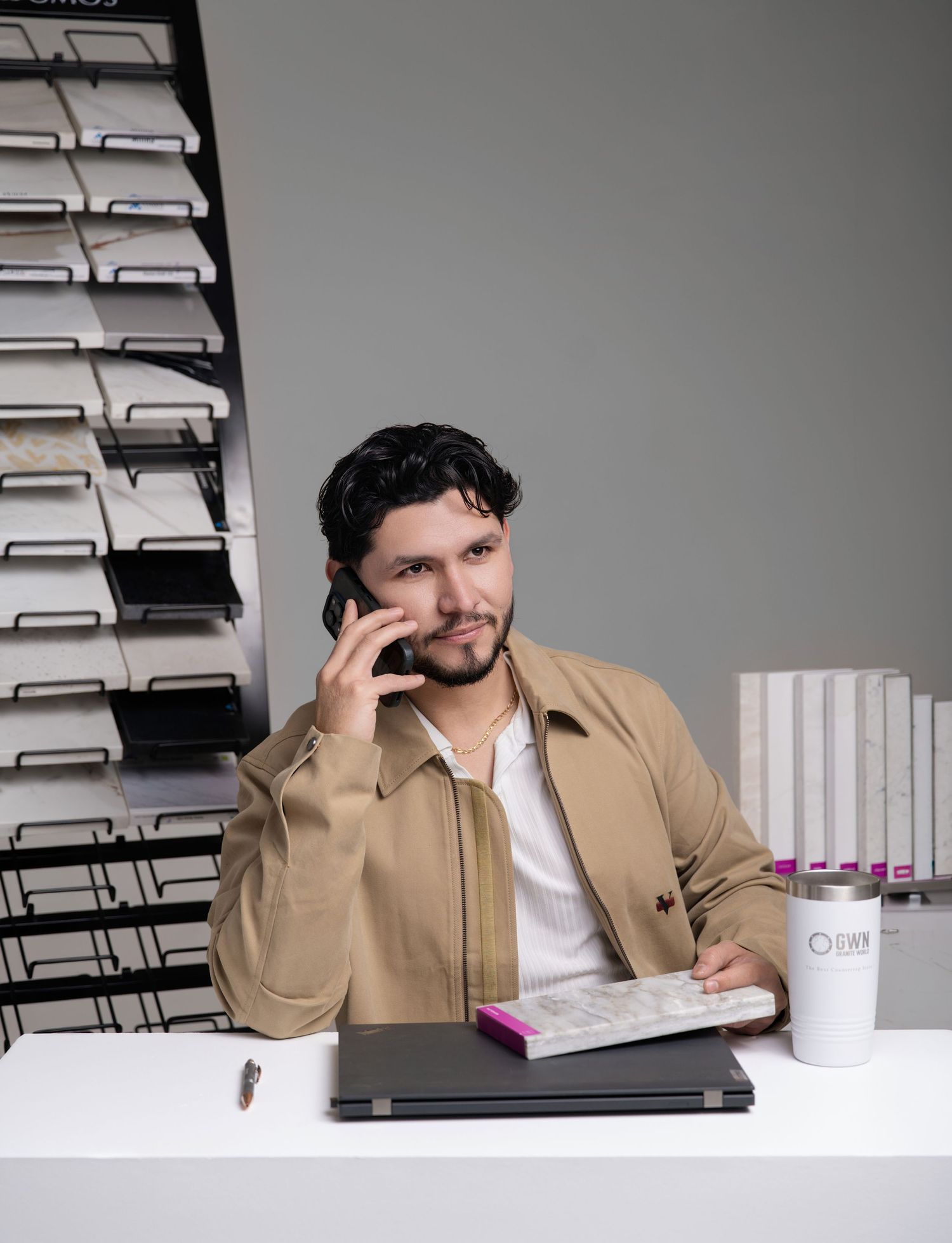 Man in beige jacket on phone call at desk with laptop, books, and tile sample display in background.