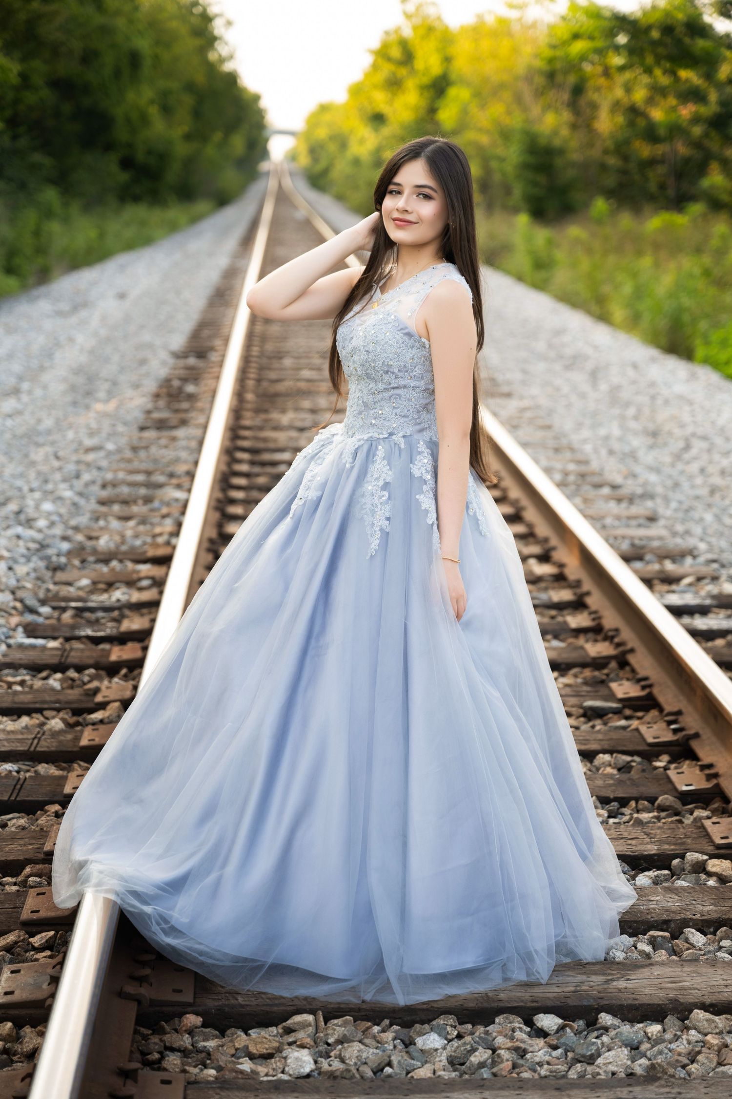 Young woman in a light blue ball gown posing elegantly on railroad tracks surrounded by lush greenery.