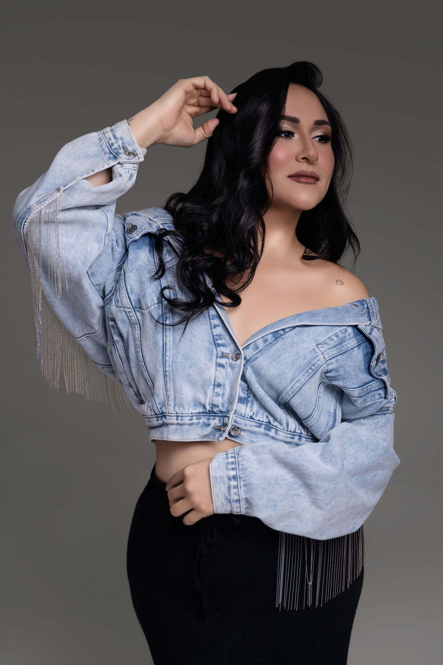 Woman with long wavy black hair poses in a cropped off-shoulder denim jacket with silver fringe and black pants against a gray backdrop.