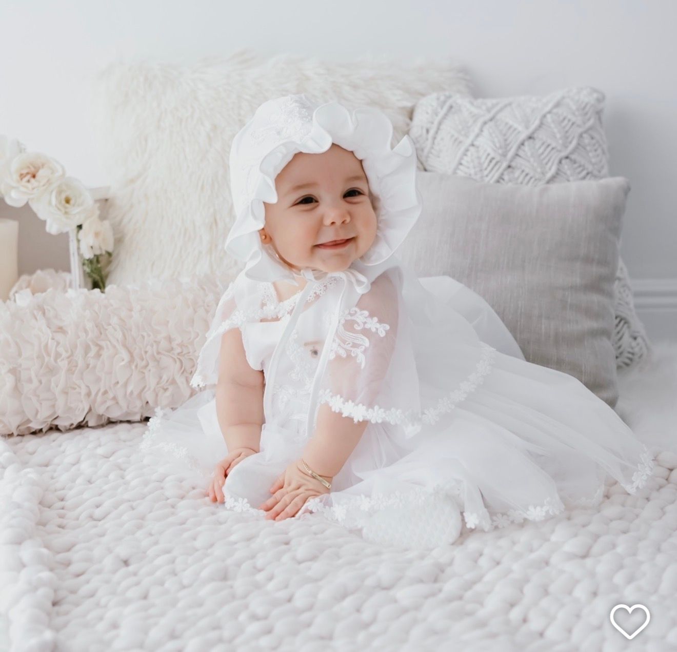 An adorable baby girl sits on a bed wearing a white lace christening gown and matching bonnet.