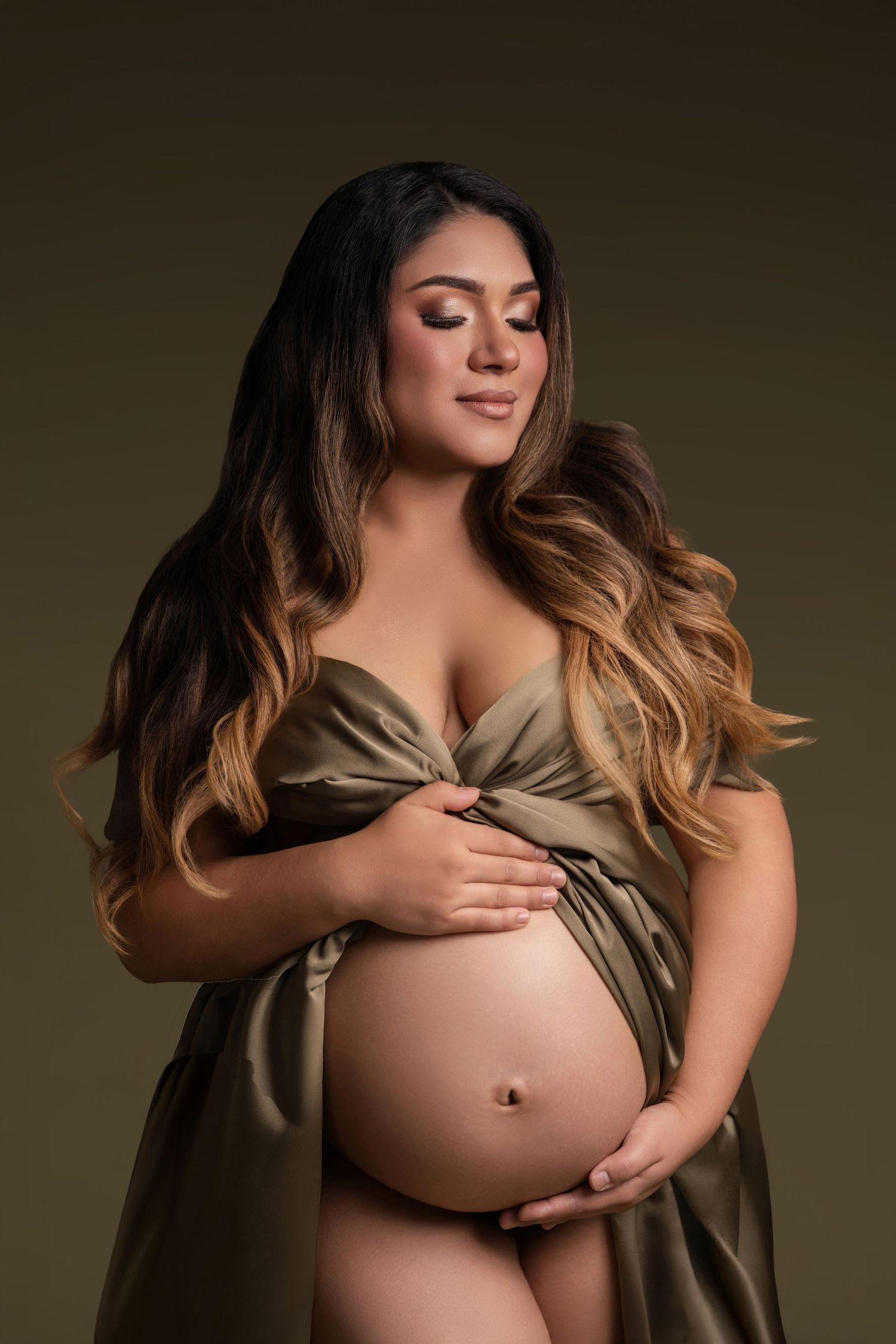 Pregnant woman in olive satin wrap gown cradling baby bump in elegant maternity studio portrait.