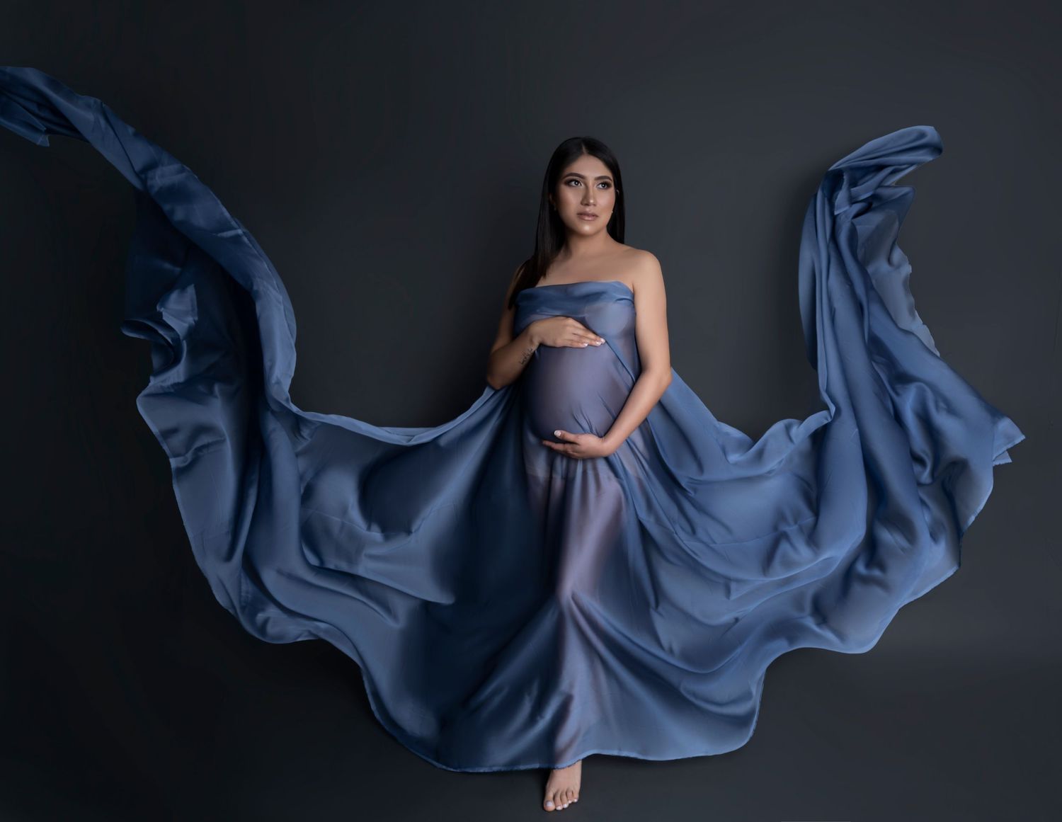 Pregnant woman in flowing blue maternity gown with dramatic fabric movement against dark studio backdrop.