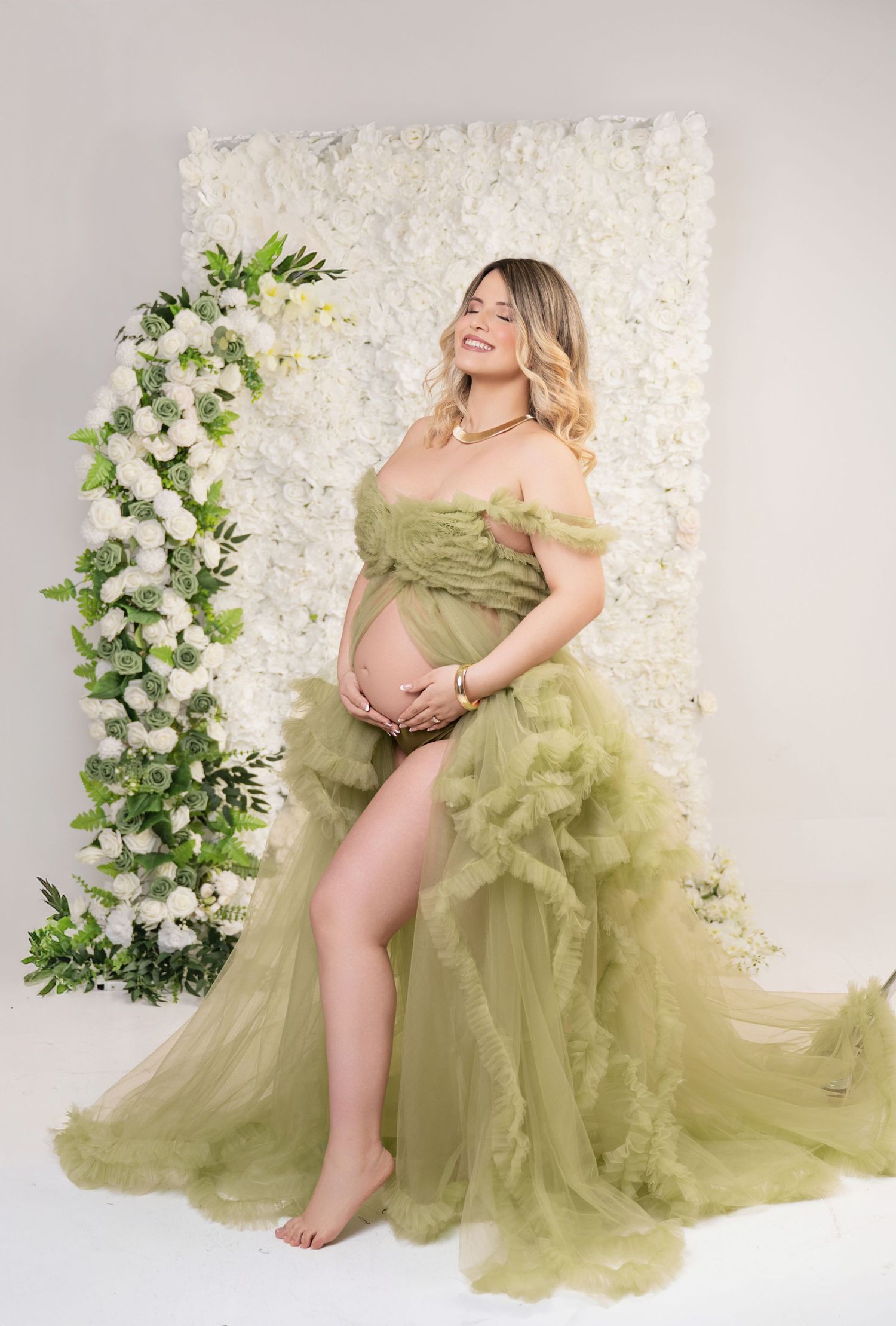 Pregnant woman in olive green tulle gown poses barefoot before white floral wall backdrop in maternity photo session.
