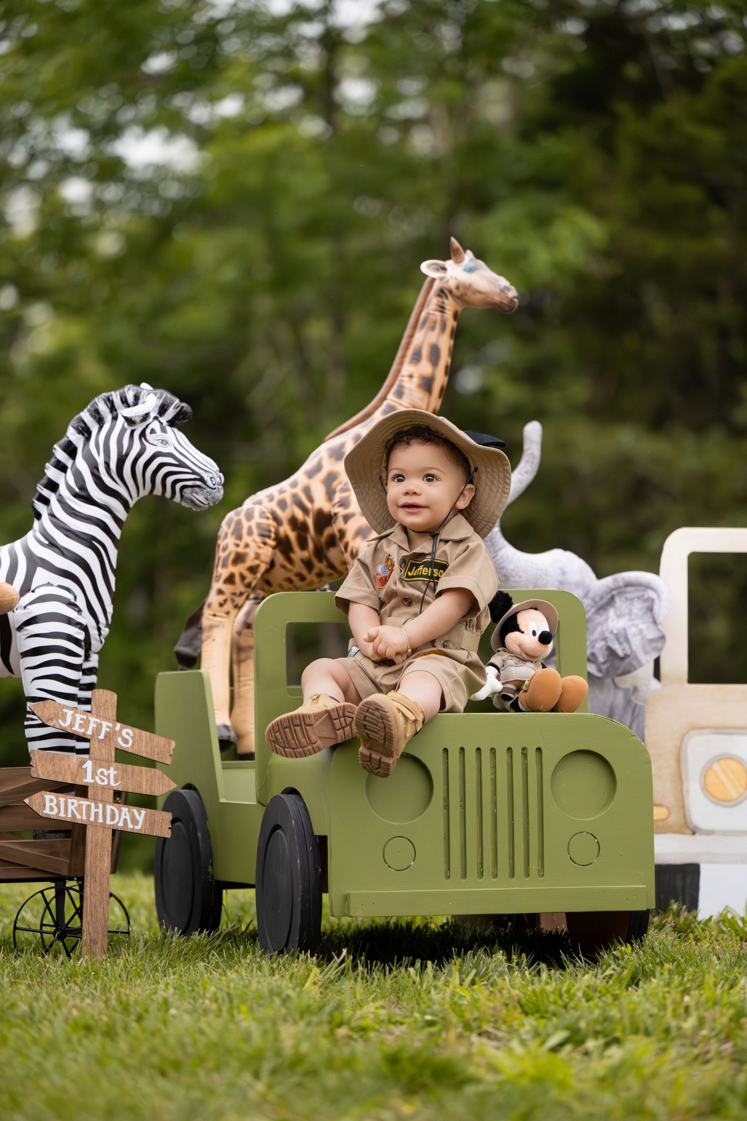 Baby in safari outfit sits in toy jeep surrounded by giraffe, zebra, and elephant figures at 1st birthday party.