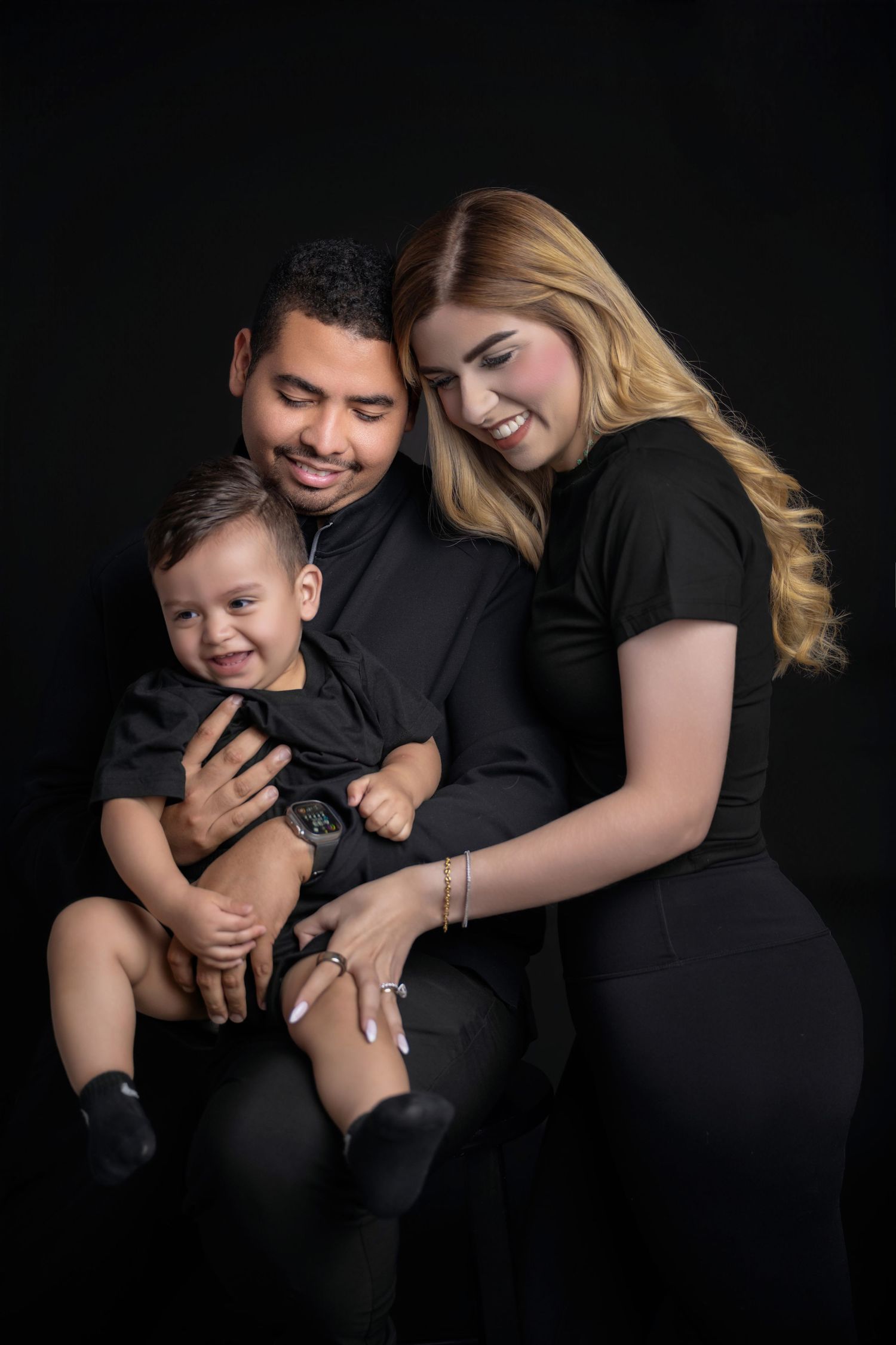 Family portrait of parents in black outfits smiling at their happy toddler son against dark background.