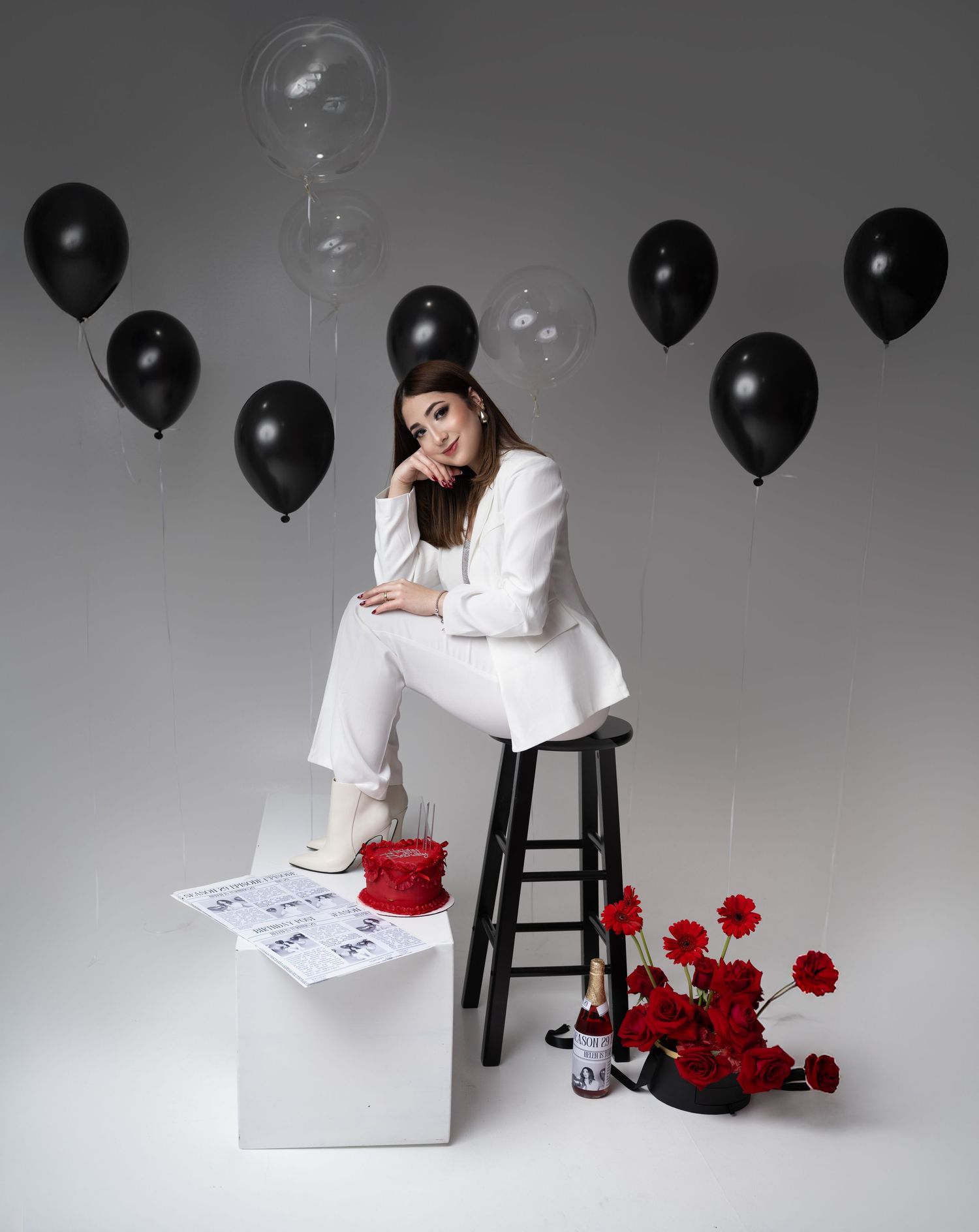 Artistic studio photo with black balloons, white outfit, red roses and scattered papers creating a moody atmosphere.