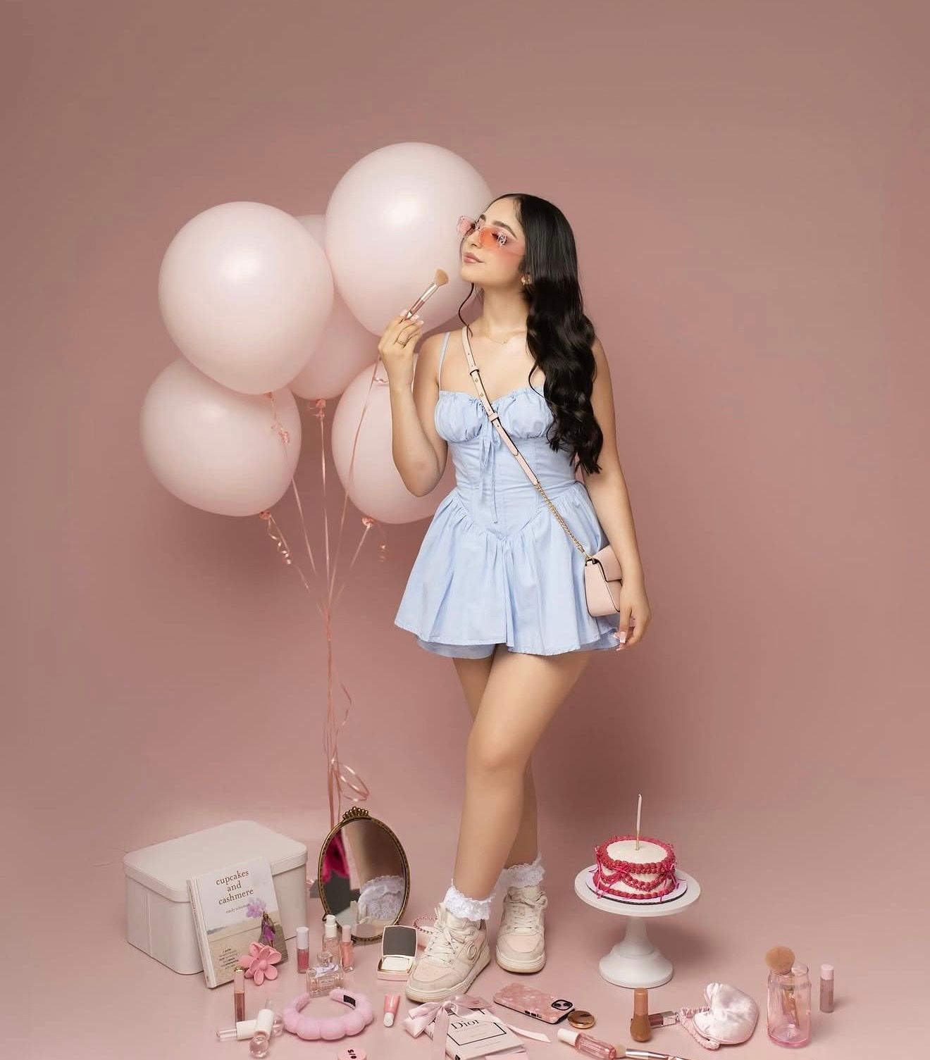 Young woman in blue mini dress poses with pink balloons, cake, and accessories on a pink background.