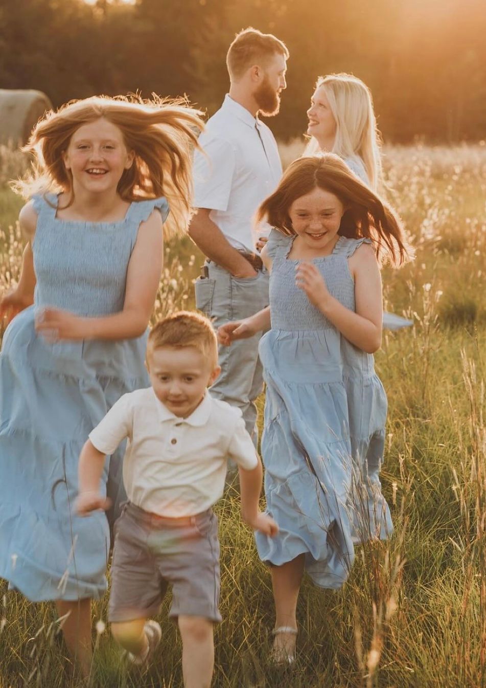 A family in coordinated blue and white outfits dances together in a sunlit meadow during golden hour.