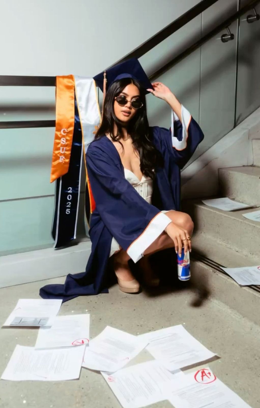 Graduate in navy cap and gown sits on stairs with scattered papers and holds a drink.