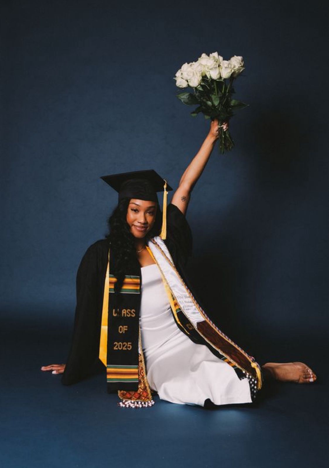 A graduate in cap and gown sits on the floor holding white roses against a dark blue backdrop during a graduation photoshoot.