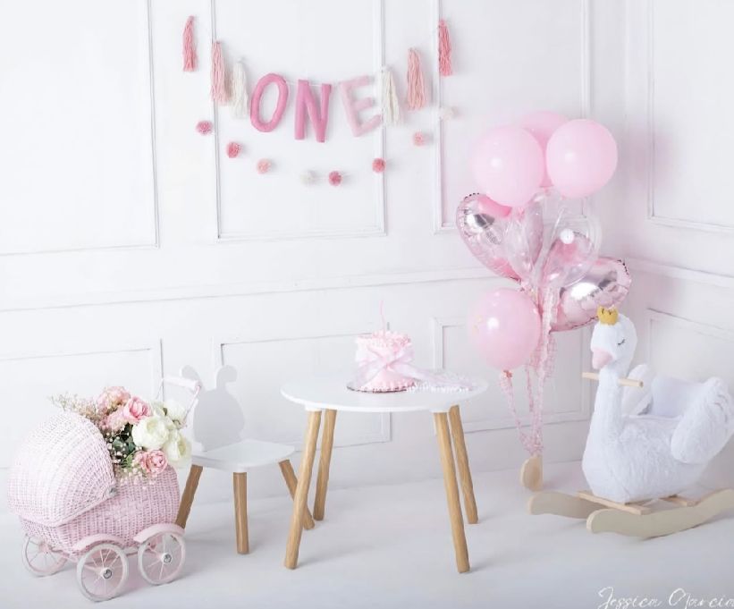 Pink baby nursery corner with rocking horse, balloons, small table and decorative pram in soft pastel colors.