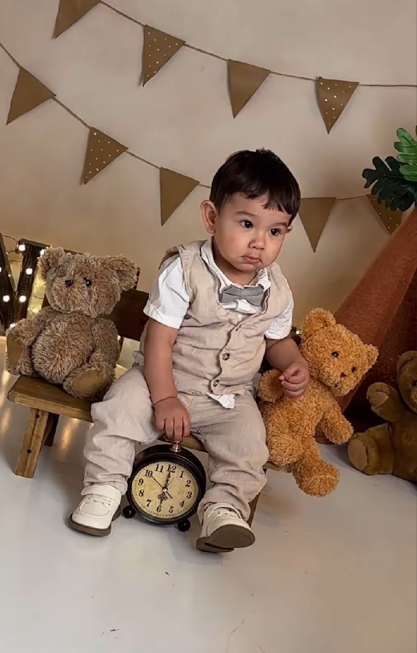 A small child in beige overalls sits with teddy bears under gold bunting decorations during a photo session.