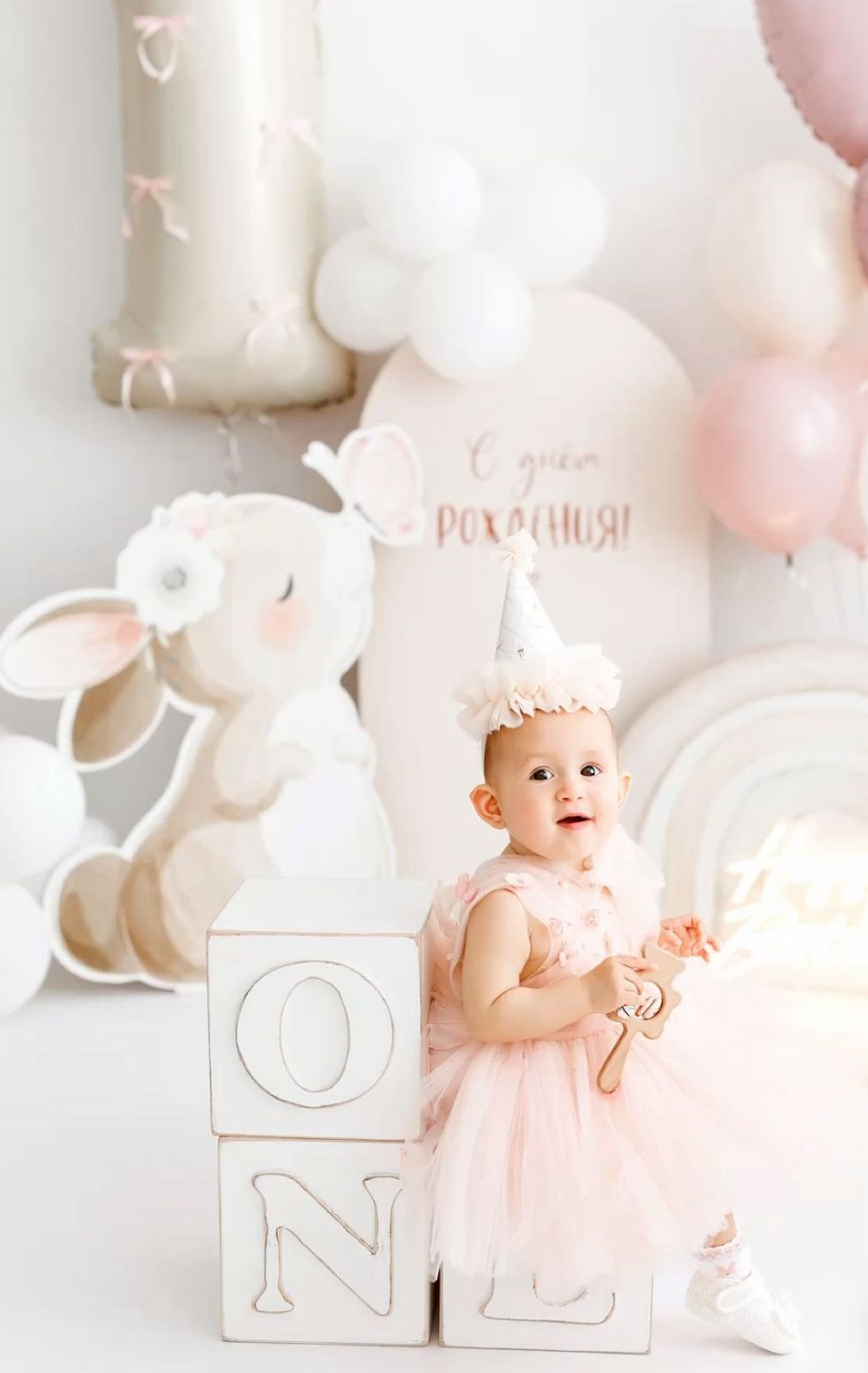 Soft pink and white first birthday party setup with balloons, plush bunny, and decorative blocks in dreamy pastel tones.